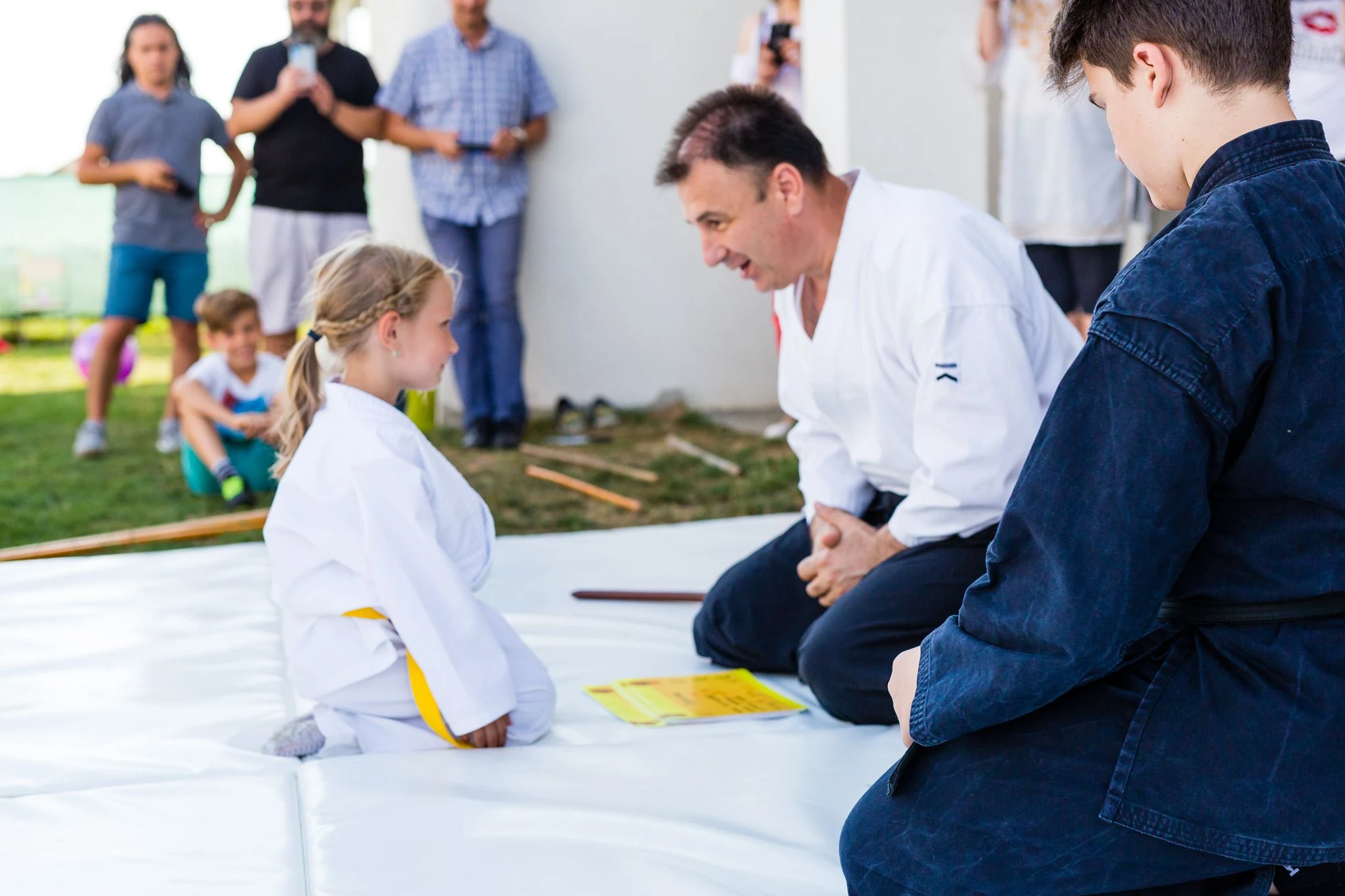 Copil în kimono alb cu centură galbenă, așezat în seiza pe saltea, ascultând instructorul de Aikido; în plan îndepărtat, părinți care privesc de pe margine.