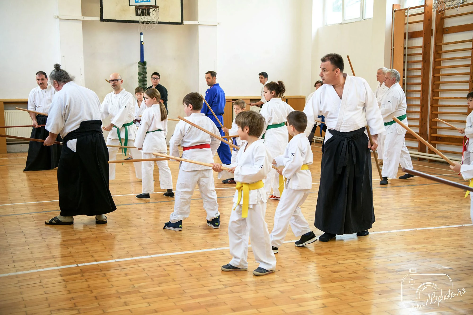 Grupă mixtă de copii și adulți în kimono alb, exersând cu bastoane de lemn într-o sală de sport, îndrumați de doi instructori de Aikido în hakama neagră.