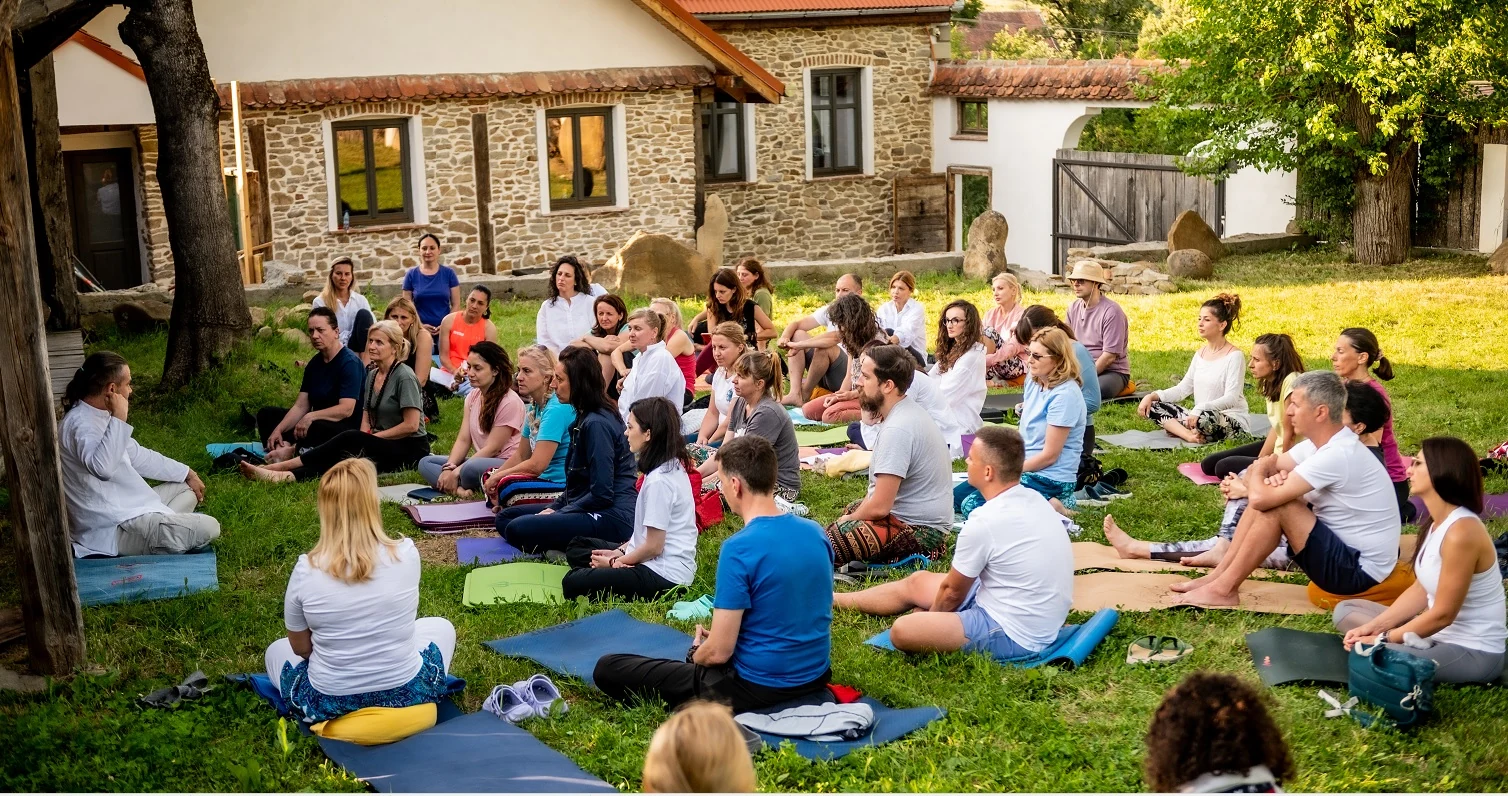 Grup de adulți așezați pe saltele pe iarbă, în fața unei case tradiționale de piatră, ascultând profesorul la o sesiune de Hatha Yoga SEVA în aer liber.