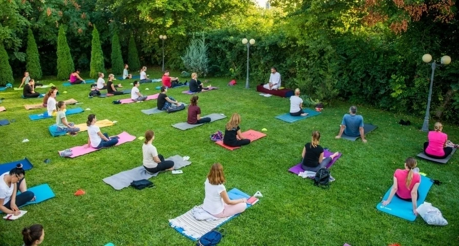 Vedere de ansamblu a unei sesiuni de Hatha Yoga SEVA în parc: zeci de participanți adulți, așezați pe saltele la distanță unul de altul, printre brazi și felinare.