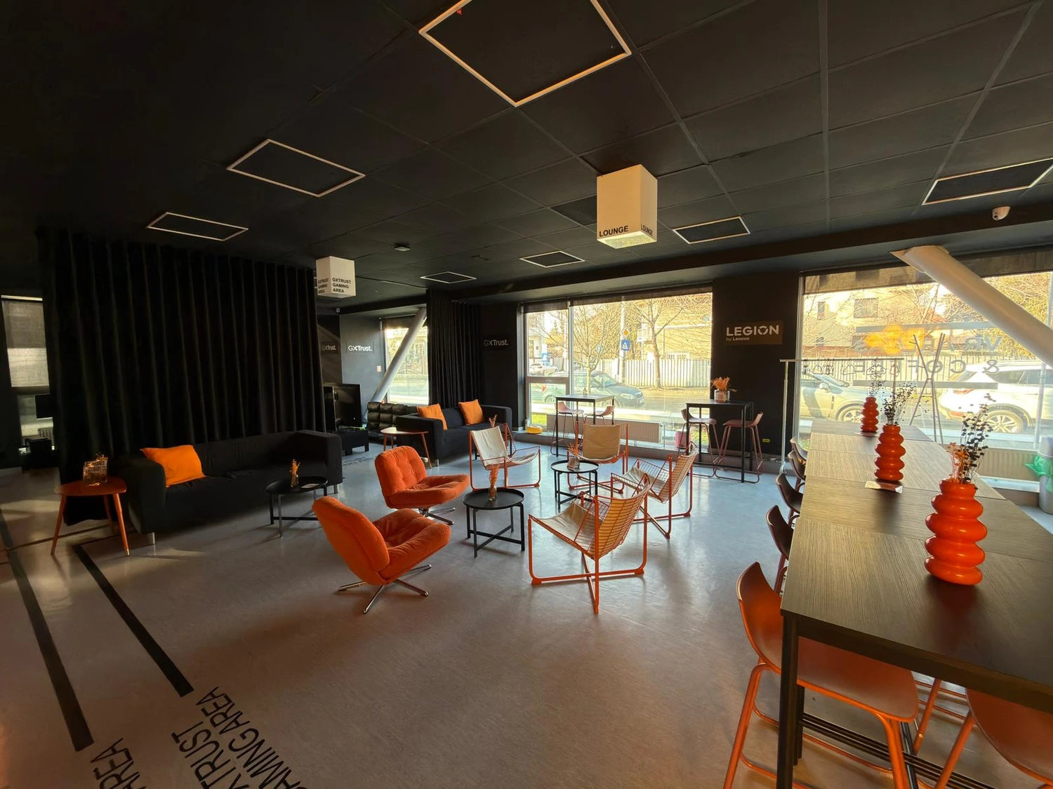 Aceeași sală în configurație de lounge: canapele și fotolii portocalii, măsuțe joase și o masă comună lungă lângă ferestrele spre stradă.